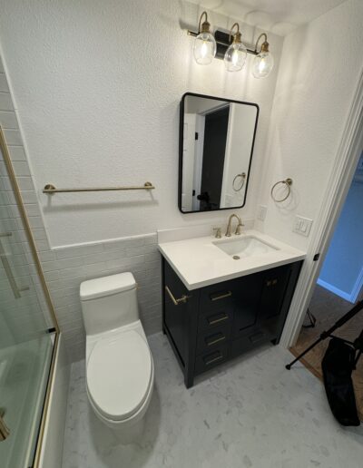 Bathroom remodel featuring a dark vanity with gold hardware, a white countertop, and a rectangular mirror with black trim. The bathroom has a modern toilet, a gold towel bar, and three clear globe lights over the mirror. The walls are white with a subtle texture, and the floor is covered with light hexagonal tiles.