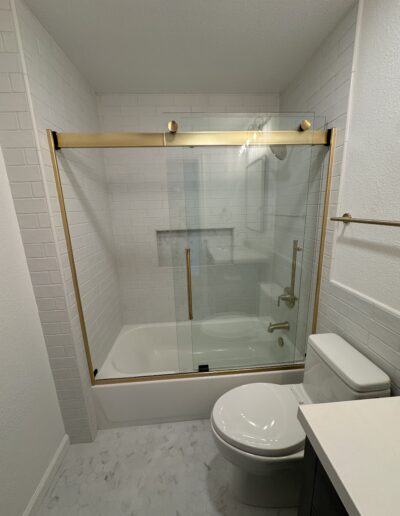 Remodeled bathroom showing a white bathtub with sliding glass doors framed in gold. The shower area is tiled with white subway tiles and includes a built-in niche. The bathroom floor is covered with light gray hexagonal tiles, and the gold hardware ties in with the modern and elegant design